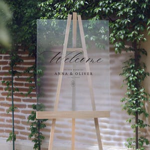 May include: A clear acrylic wedding welcome sign on a wooden easel. The sign features elegant script that reads "Welcome" above the names "Anna & Oliver" and the wedding date. The background includes greenery and a brick wall.