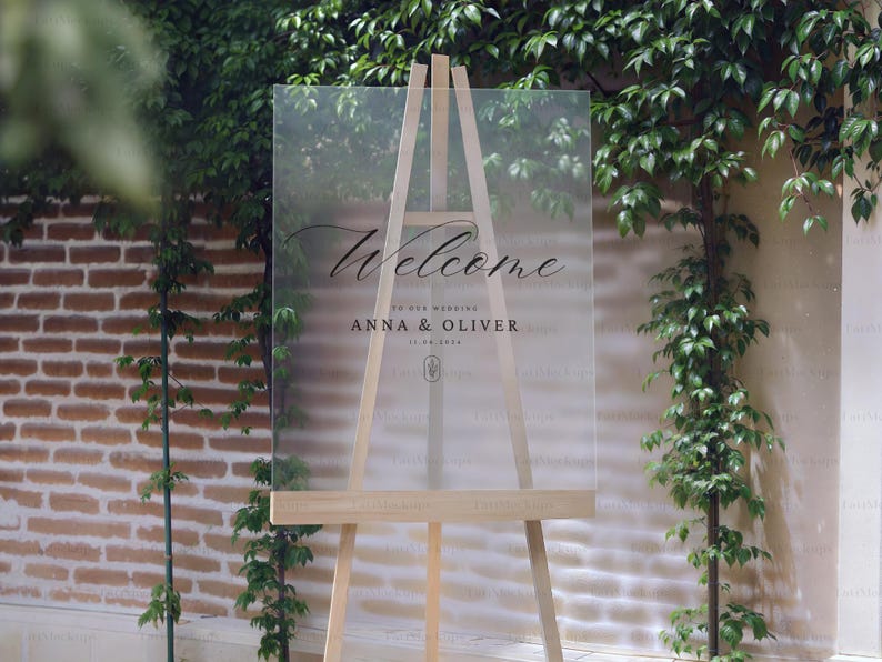 May include: A clear acrylic wedding welcome sign on a wooden easel. The sign features elegant script that reads "Welcome" above the names "Anna & Oliver" and the wedding date. The background includes greenery and a brick wall.