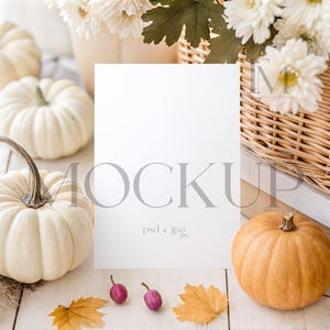 Könnte beinhalten: Eine Mockup-Szene mit weißen Kürbissen, einem kleinen orangefarbenen Kürbis und einer leeren weißen Karte. Die Szene umfasst einen Weidenkorb mit weißen Blumen, Herbstblättern und kleinen lila Beeren. Das Wort "MOCKUP" ist auf die Karte gedruckt.