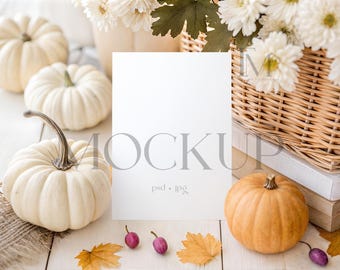 May include: A mockup scene featuring white pumpkins, a small orange pumpkin, and a blank white card. The scene includes a woven basket with white flowers, fall leaves, and small purple berries. The word "MOCKUP" is printed on the card.