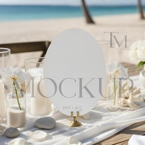 Op de afbeelding: Een tafelsetting met strandthema met een blanco wit ovaal bord op een gouden standaard. De tafel is versierd met kaarsen, bloemen en decoratieve elementen. De achtergrond toont de oceaan en een zandstrand.