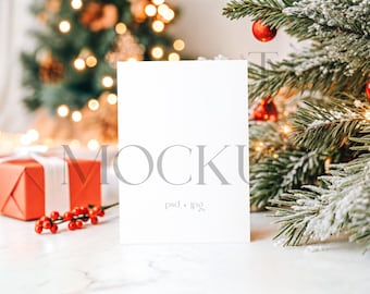 May include: A blank white card mockup with the word "MOCKUP" in light gray. A red gift box with a white ribbon, red berries, and a Christmas tree with red ornaments and lights are in the background. The text "psd + jpg" is below the word "MOCKUP".