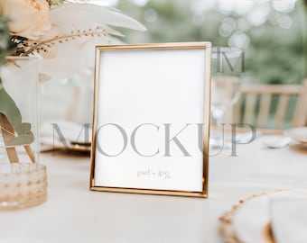 May include: A gold-framed picture frame with a white interior stands on a white tablecloth. The frame is rectangular and has a thin, gold border. The background is blurred, with hints of flowers and greenery, suggesting a wedding or event setting.
