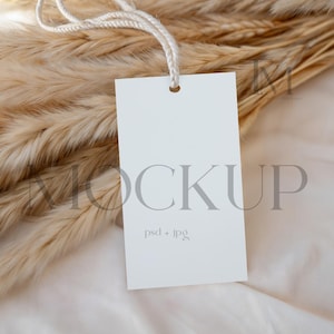 Può includere: Un'etichetta rettangolare bianca vuota con un cordino, appoggiata su un letto di erba della pampa beige essiccata. L'etichetta presenta la parola "MOCKUP" in lettere grigie e il testo "psd + jpg" in un carattere più piccolo.