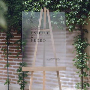 May include: A clear acrylic wedding welcome sign on a wooden easel. The sign features the names "ESTELLE & PEDRO" in elegant black font, with the text "WELCOME TO THE WEDDING OF" and "THANK YOU FOR CELEBRATING WITH US!" also displayed.