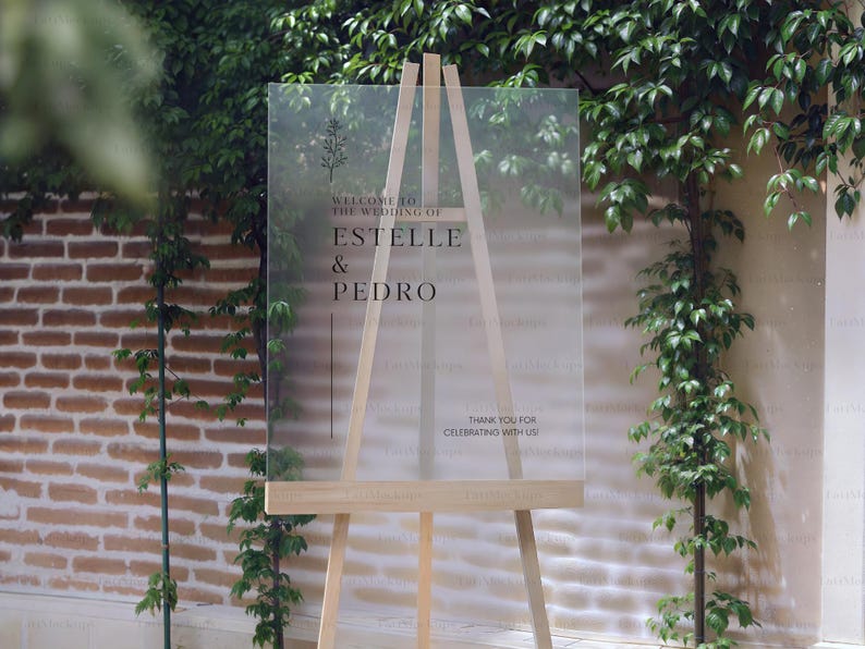 May include: A clear acrylic wedding welcome sign on a wooden easel. The sign features the names "ESTELLE & PEDRO" in elegant black font, with the text "WELCOME TO THE WEDDING OF" and "THANK YOU FOR CELEBRATING WITH US!" also displayed.