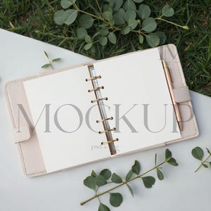Puede incluir: Una agenda rosa claro abierta con un bolígrafo dorado, con la palabra "MOCKUP" en las páginas. La agenda está sobre una superficie blanca con follaje verde y hierba en el fondo. La agenda está abierta a páginas en blanco.