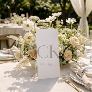 May include: A vertical white rectangular menu card with the text MOCKUP and psd + jpg printed on the front, positioned on a table set for a formal event with a floral centerpiece of white roses, daisies, and greenery, plates, and glassware.