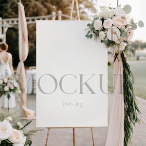May include: A blank white sign on a gold easel, with the word "MOCKUP" in gray. The sign is decorated with a floral arrangement of white and pink roses. The background includes a blurred outdoor setting.