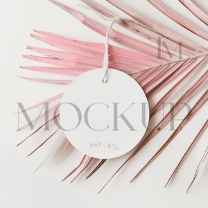 May include: A white round tag with the word "MOCKUP" printed on it, hanging from a pink palm leaf. The tag has the text "psd + jpg" printed on it.