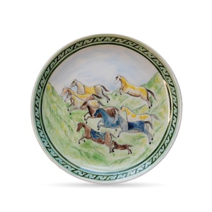 Puede incluir: Un plato redondo de cerámica con una escena pintada a mano de caballos corriendo por un paisaje verde y azul. El plato tiene un borde verde con dibujos. Los caballos son de color marrón, amarillo y azul.