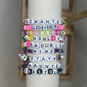 Pu&ograve; includere: Una pila di braccialetti di perline con perline di lettere bianche che compongono frasi come "ENCHANTED", "LOVER" e "FEARLESS". I braccialetti presentano perline colorate in rosa, giallo e viola.