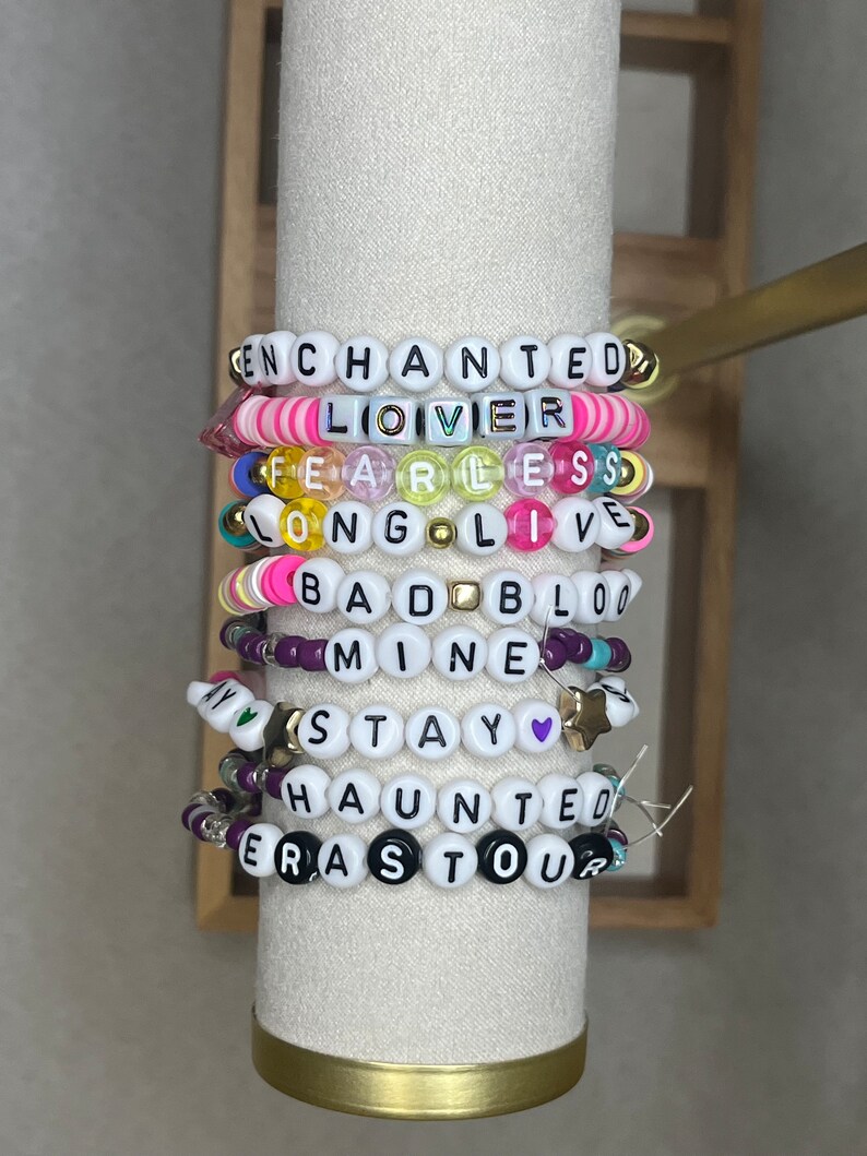 Pu&ograve; includere: Una pila di braccialetti di perline con perline di lettere bianche che compongono frasi come "ENCHANTED", "LOVER" e "FEARLESS". I braccialetti presentano perline colorate in rosa, giallo e viola.