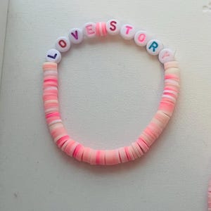 Pu&ograve; includere: Un braccialetto di perline rosa e bianche con le parole "LOVE STORY" scritte con perline colorate. Il braccialetto &egrave; fatto di perline piatte a forma di disco in tonalit&agrave; di rosa e bianco.