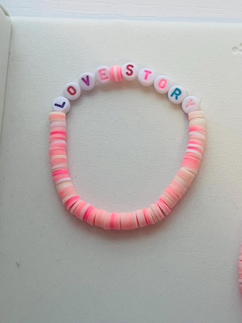 Pu&ograve; includere: Un braccialetto di perline rosa e bianche con le parole "LOVE STORY" scritte con perline colorate. Il braccialetto &egrave; fatto di perline piatte a forma di disco in tonalit&agrave; di rosa e bianco.