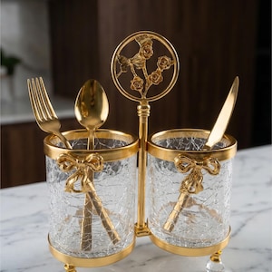 May include: A gold-toned, double utensil holder with clear, crackled glass containers. The holder features a decorative floral design and bow accents. Includes a fork, spoon, and knife. The holder is supported by clear, crystal-like feet.