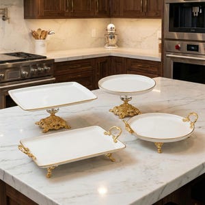 May include: Four white serving platters with gold trim and ornate gold bases and handles are displayed on a marble countertop. The platters are in various shapes, including rectangular and round, and are set against a kitchen backdrop.
