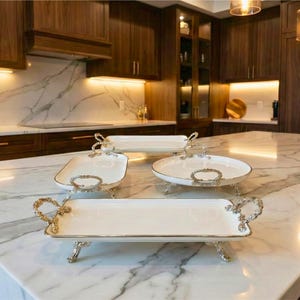 May include: Four white serving trays with gold-colored accents and ornate handles are displayed on a marble countertop. The trays are in various shapes: rectangular, square, and oval. The kitchen features dark wood cabinetry and marble backsplash.