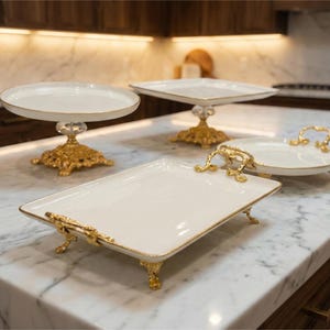 May include: Four elegant serving platters and cake stands with white surfaces and gold accents are displayed on a marble countertop. The stands have ornate gold bases and handles, showcasing a luxurious aesthetic.