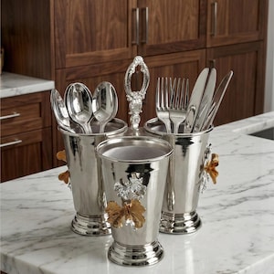 May include: Silverware caddy with three compartments, holding spoons, forks, and knives. The caddy has a decorative handle and is adorned with gold and silver floral accents. It sits on a marble countertop in front of a wooden cabinet.
