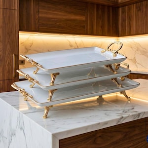 May include: A three-tiered serving tray set. Each white rectangular tray has gold trim and ornate gold handles and legs. The trays are stacked on a marble countertop in a kitchen setting.