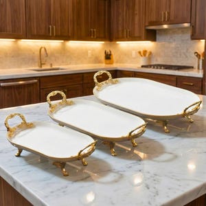 May include: Three white serving trays with gold-colored trim and handles are displayed on a marble countertop. The trays are oval-shaped and rest on small, decorative feet. The kitchen background features wooden cabinets and a stainless steel oven.