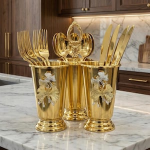May include: A gold-colored flatware holder with three compartments, each holding forks, spoons, and knives. The holder is decorated with a silver and gold floral design. The set is displayed on a marble countertop in a kitchen setting.