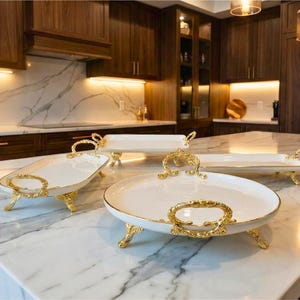 May include: A collection of white serving trays with gold-colored handles and feet, displayed on a marble countertop. The trays vary in shape and size, with a round tray in the foreground. The kitchen features dark wood cabinetry.