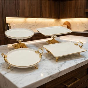 May include: Four white serving platters with gold trim and ornate gold bases and handles are displayed on a marble countertop. The platters are in various shapes, including round and rectangular, and are arranged in a kitchen setting.