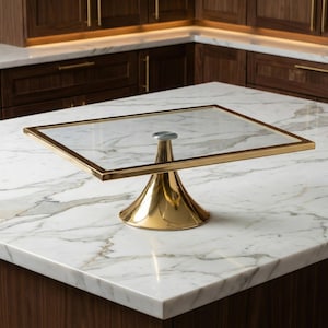 May include: A gold-toned metal cake stand with a square glass top. The stand has a flared base and a central support. The countertop is white marble with gray veining. The background features dark wood cabinetry.