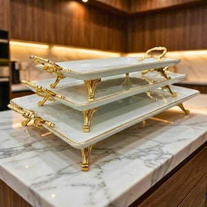 May include: A three-tiered serving tray with a white surface and gold trim. The tray has gold handles and ornate gold legs. The trays are stacked on top of each other, creating a tiered effect. The trays are on a marble countertop.