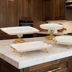May include: A collection of white ceramic serving platters and cake stands with gold-colored ornate bases and accents. The platters are rectangular and round, with some featuring handles. The items are displayed on a marble countertop.