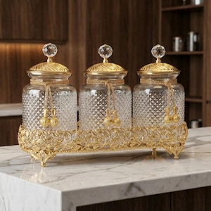 Peut inclure: Trois bocaux en verre avec des couvercles dorés et des bases dorées ornées. Chaque bocal présente un motif en losange et un bouton en cristal. Les bocaux sont posés sur un comptoir en marbre, dans une cuisine.