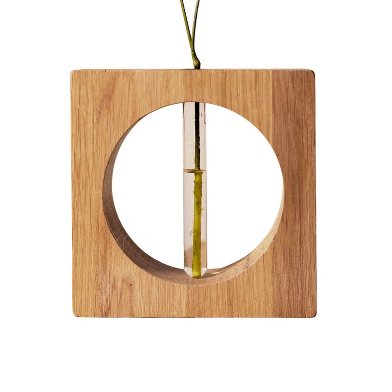 May include: A square wooden vase with a circular cutout. A clear glass tube is suspended in the center, holding a single stem. The wood has a natural grain and a light brown color. The vase is designed for a minimalist aesthetic.