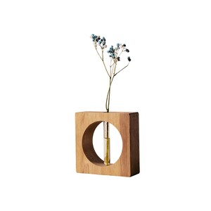 May include: A square wooden vase with a circular cutout, holding a small glass tube with a single stem of blue and white flowers. The vase is made of light brown wood and has a minimalist design.