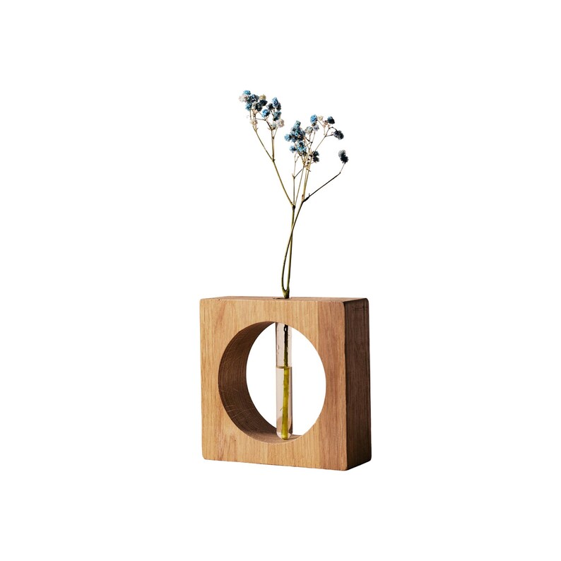 May include: A square wooden vase with a circular cutout, holding a small glass tube with a single stem of blue and white flowers. The vase is made of light brown wood and has a minimalist design.