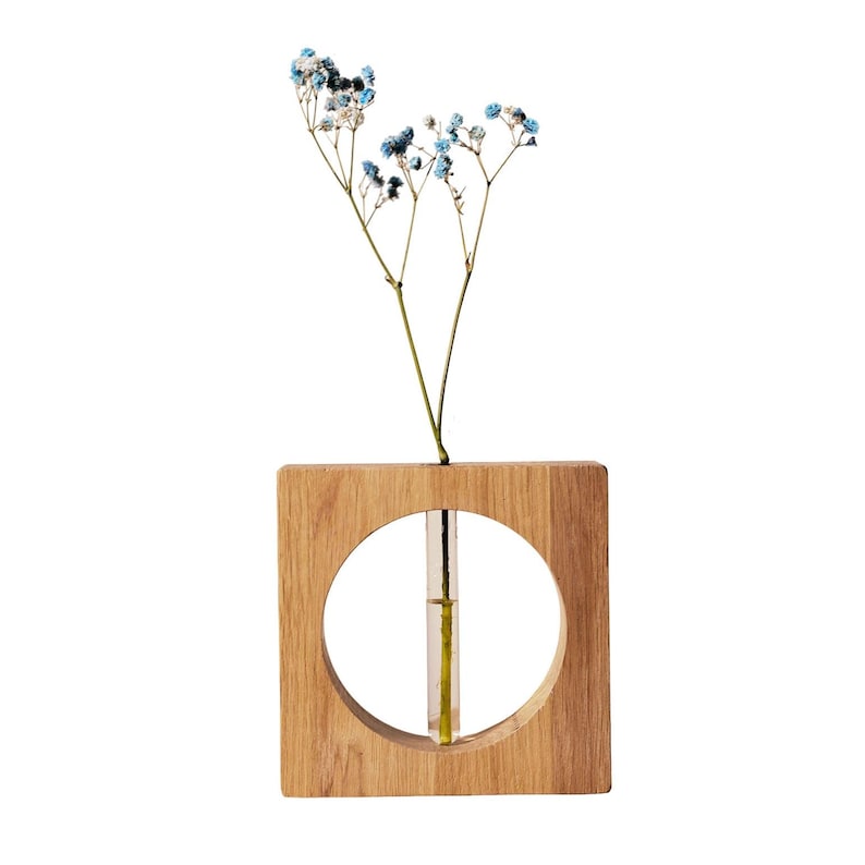 May include: A square wooden vase with a circular cutout, holding a sprig of blue and white flowers. The vase has a natural wood grain finish and a small glass tube inside to hold water for the flowers.