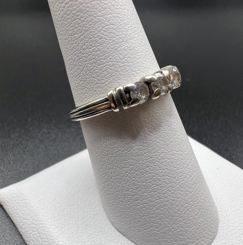 Op de afbeelding: Een zilveren ring met vijf heldere stenen in een rij. De ring ligt op een witte ringstandaard.