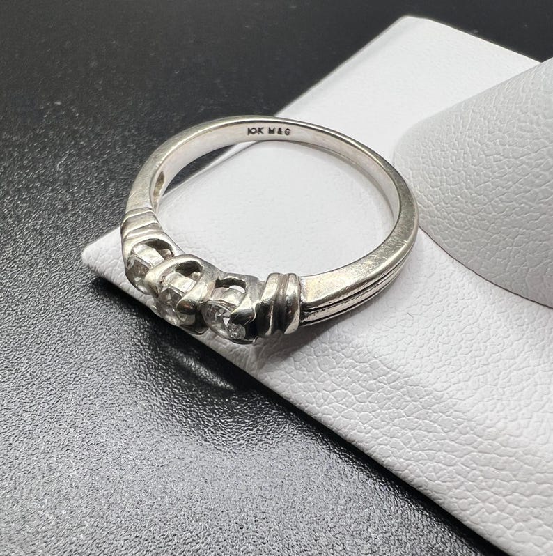 Op de afbeelding: Een zilveren bandring met drie kleine diamanten gezet in een decoratief ontwerp. De ring is gemarkeerd met "10K MAG" aan de binnenkant.