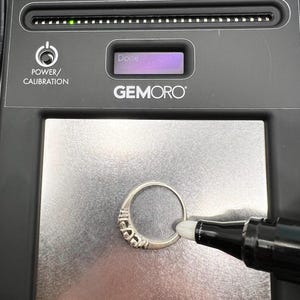 Op de afbeelding: Een zwarte Gemoro Au'racle Analyzer met een zilveren ring op het testoppervlak. Het apparaat heeft een digitaal display dat "Klaar" aangeeft en een rij lampjes die de karaat-zuiverheid van het metaal aangeven. Het apparaat wordt gebruikt om de karaat-zuiverheid van gouden en platina sieraden te testen.