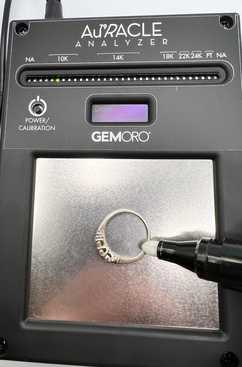 Op de afbeelding: Een zwarte Gemoro Au'racle Analyzer met een zilveren ring op het testoppervlak. Het apparaat heeft een digitaal display dat "Klaar" aangeeft en een rij lampjes die de karaat-zuiverheid van het metaal aangeven. Het apparaat wordt gebruikt om de karaat-zuiverheid van gouden en platina sieraden te testen.