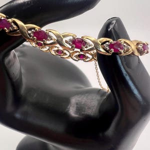 May include: A gold bracelet featuring oval-cut ruby gemstones and small diamond accents. The bracelet has an X-shaped design with a chain closure. The gemstones are a deep red color, and the metal has a polished finish.