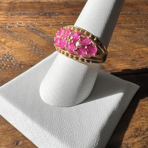 May include: A gold ring with a band of pink gemstones. The ring is set on a white display stand.
