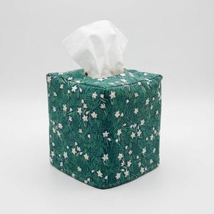 May include: A square tissue box cover with a green floral pattern. The cover is adorned with white flowers and green leaves. White tissues are pulled from the top opening. The cover is made of fabric.