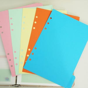 May include: A stack of colorful, hole-punched paper sheets in pink, mint green, yellow, orange, and blue. The papers are arranged in a fan shape, with the blue sheet at the front. The papers are likely for use in a binder.