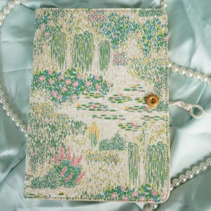 May include: A rectangular journal cover with a landscape design featuring a pond with lily pads, weeping willows, and pink flowers. The cover is secured with a wooden button and a white string. A pearl necklace is draped around the journal.