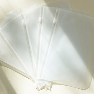 May include: A stack of clear, plastic binder pouches with white zippers and three-hole punch edges. The pouches are translucent, allowing visibility of contents. The image is lit with soft, natural light, highlighting the texture and transparency of the material.