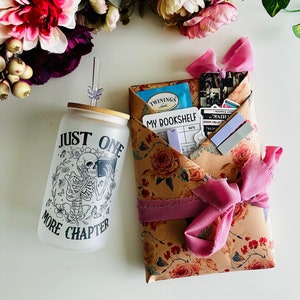 Pu&ograve; includere: Un regalo avvolto in carta marrone con un motivo floreale e un nastro rosa. Il regalo include un bicchiere di vetro trasparente con uno scheletro nero che legge un libro e il testo "Just One More Chapter". Il regalo include anche una scatola di t&egrave;, un registro dei libri e alcuni adesivi.