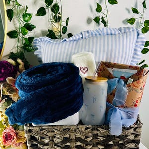 May include: A wicker basket filled with a blue throw blanket, a white pillow with blue stripes, a white sock with a red heart, a clear glass water bottle with a blue bow design, and a wrapped gift box with a blue ribbon.
