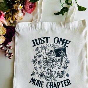 Pu&ograve; includere: Borsa di tela bianca con testo nero e un'immagine di uno scheletro che legge un libro. Il testo dice "Just One More Chapter".